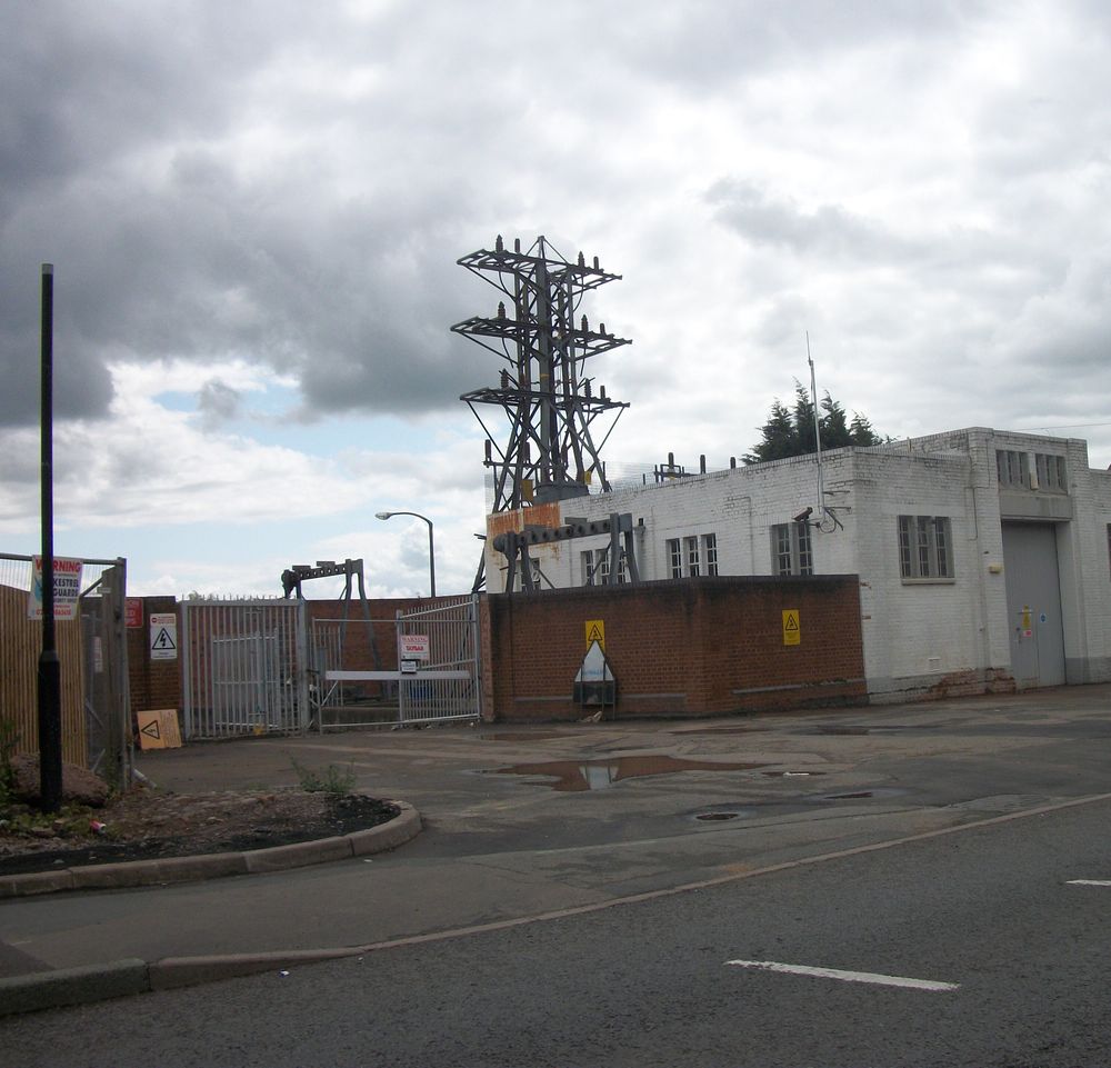An electricity substation and pylon behind a brick wall under a grey sky.